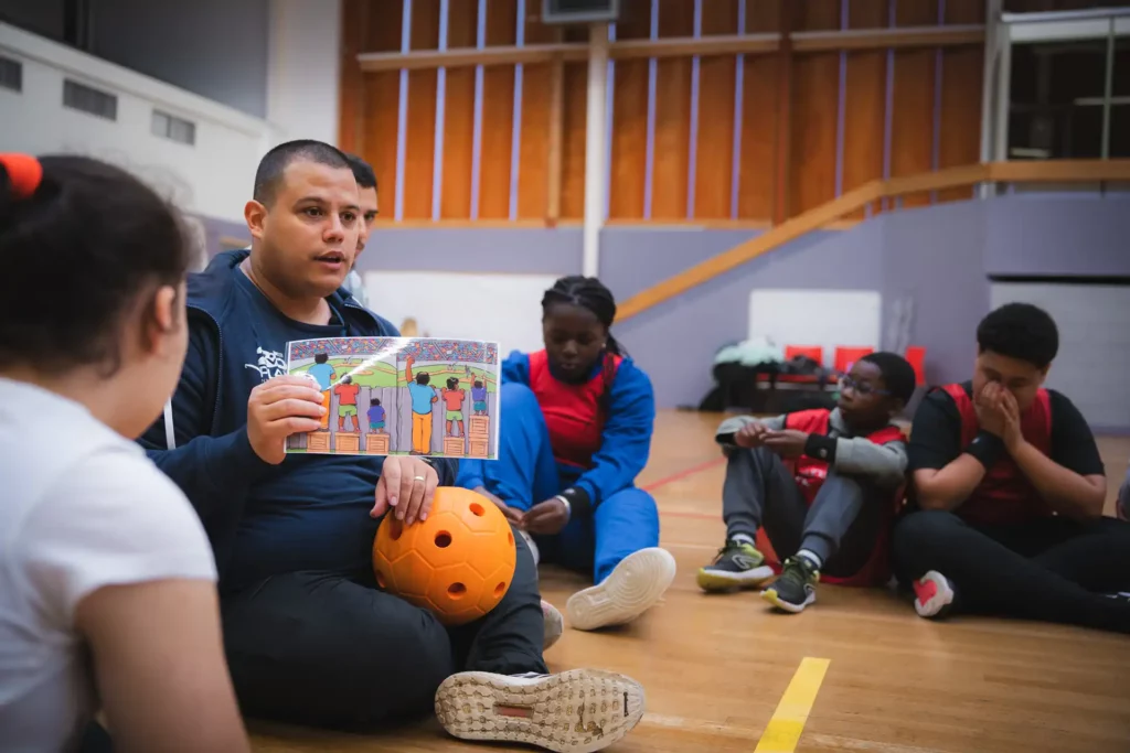 Homme éducateur qui est entouré par des enfants dans un gymnase. Il est entrain de leurs parler en s'aidant d'un livre illustré.
